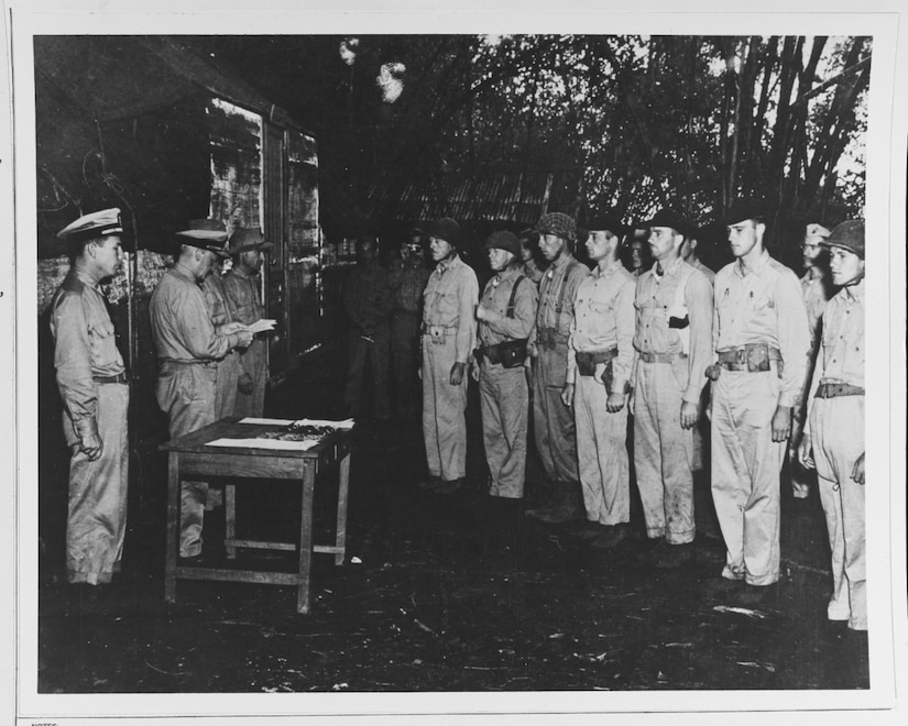 A half-dozen men stand at attention, looking toward four men standing behind a small table.