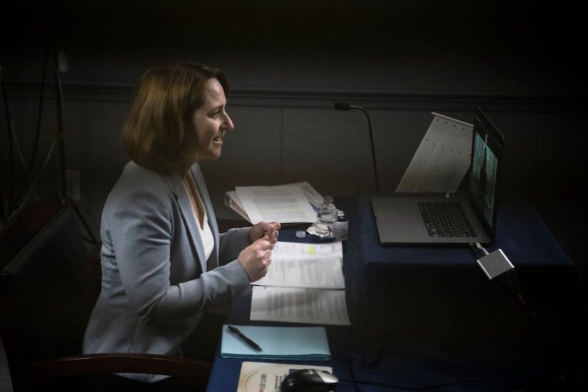 Deputy defense secretary sits at a table and speaks in front of a computer screen.