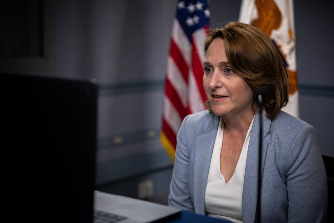 Deputy defense secretary sits at a table and speaks in front of a computer screen.