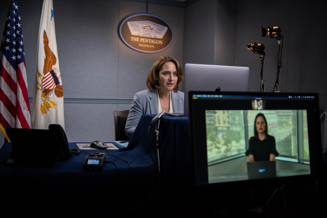 Deputy defense secretary sits at a table and speaks in front of a computer screen.