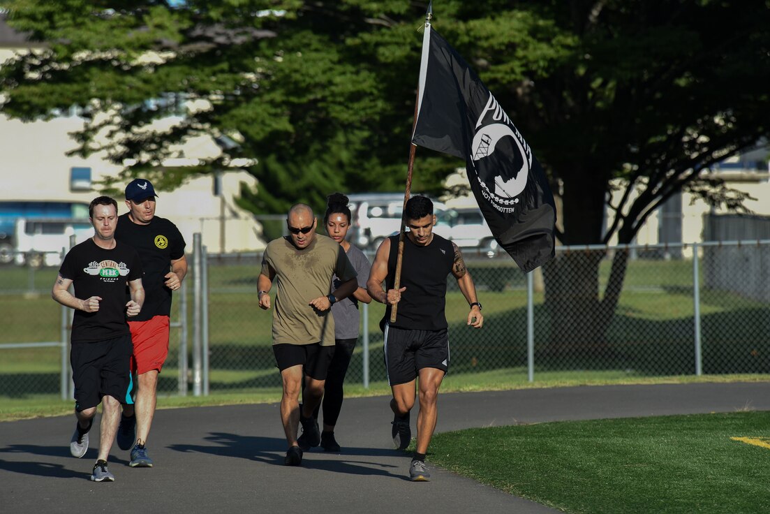 Five participants run on a track, while the right most member holds up the POW/MIA flag up above his shoulder.