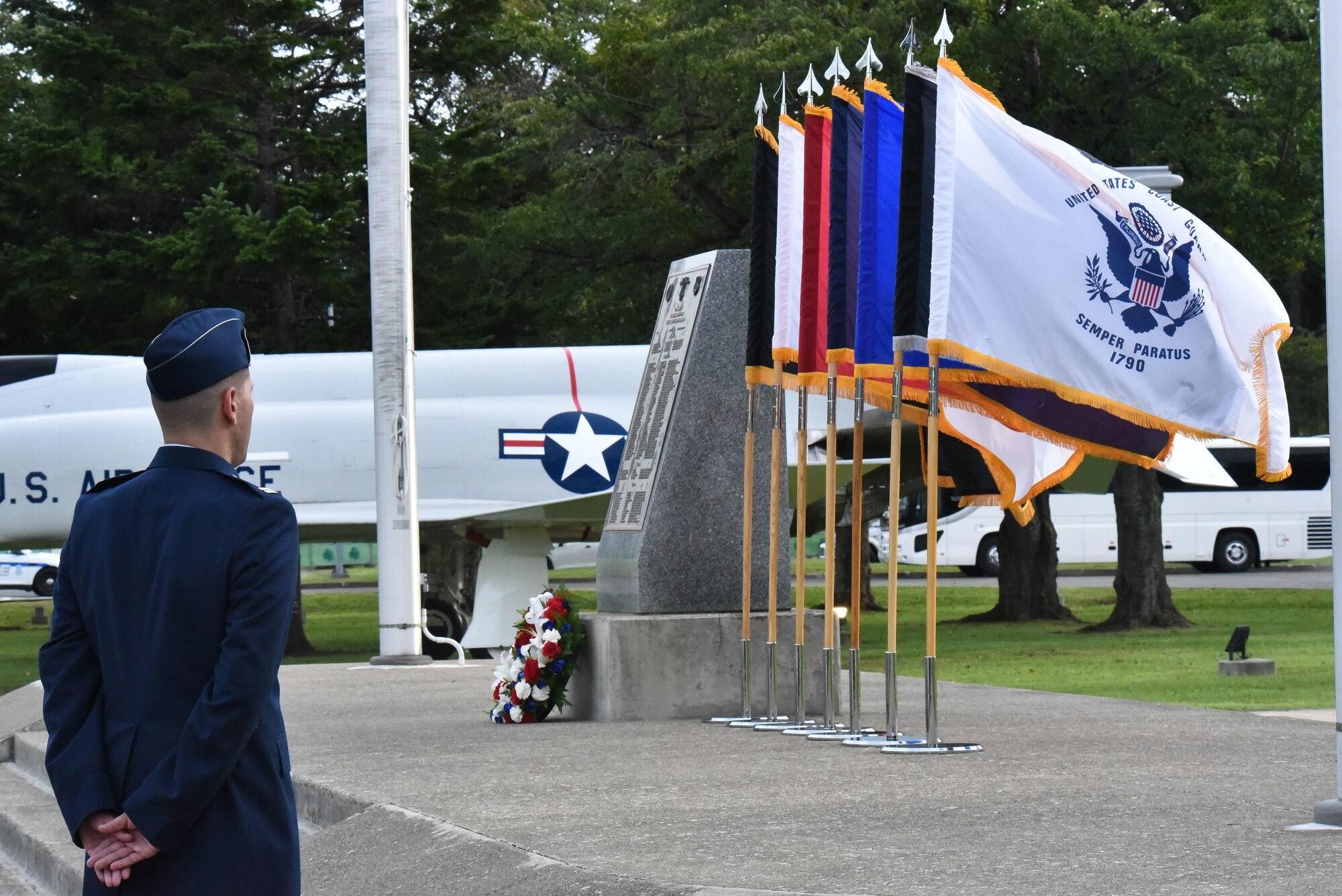 A man in uniform stands in front of flags, flowers and a memorial stone.
