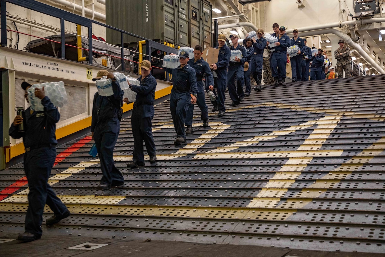 Sailors carry boxes of food and water down a ramp.
