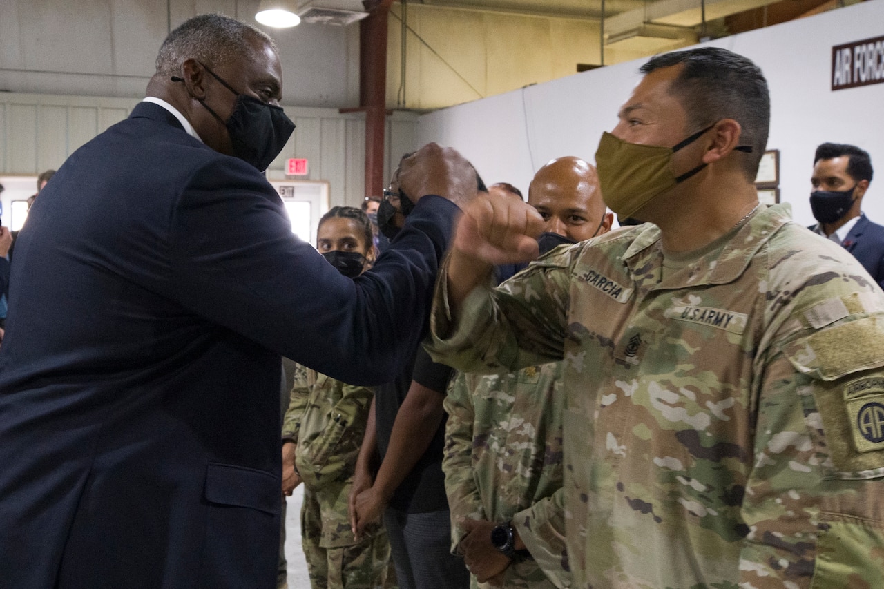 A man dressed in a business suit bumps elbows with a service member.