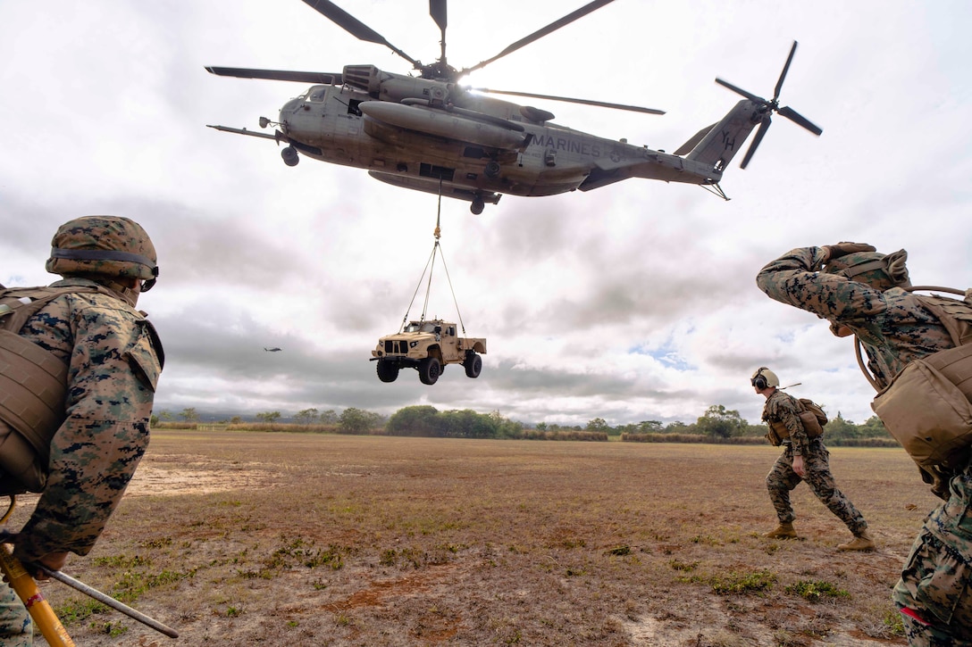 Three Marines surround an airborne helicopter that is hauling a military vehicle.