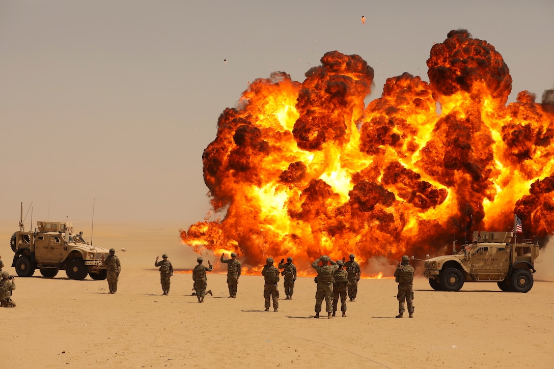 Soldiers stand near military vehicles as a large fire erupts in a sandy area.