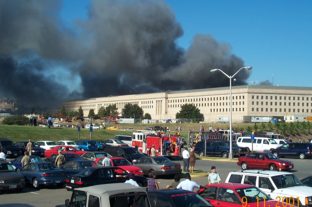 Smoke emanates from a burning building. Firetrucks are in a parking lot.