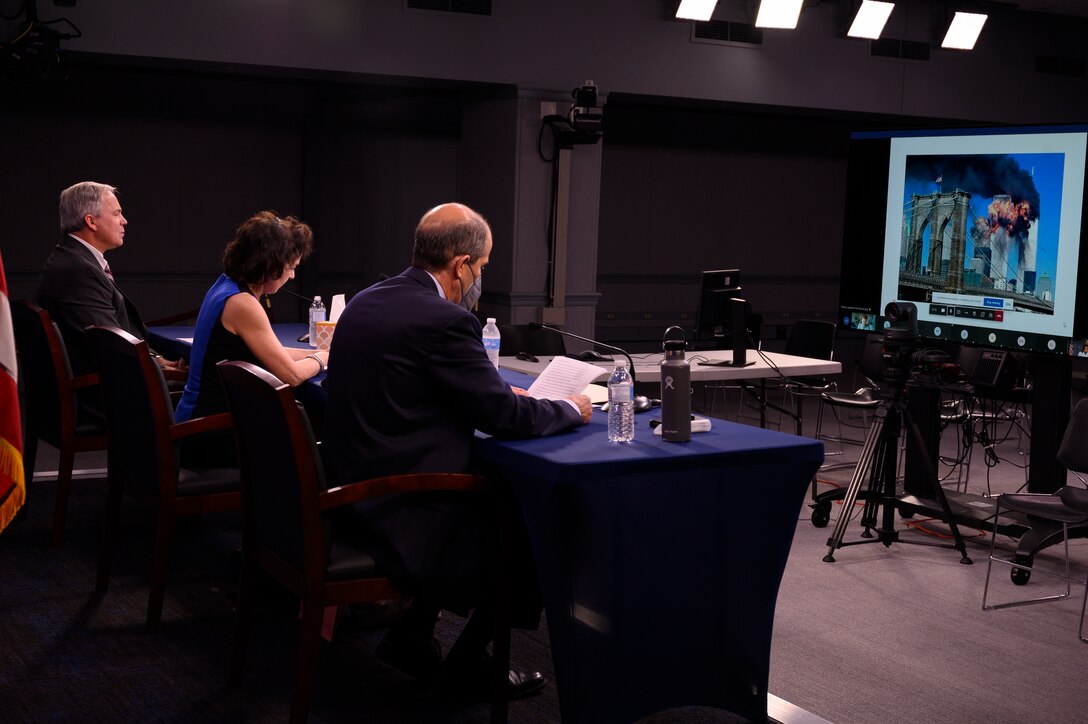 In a studio setting, three individuals sit at a table and look at a computer monitor.