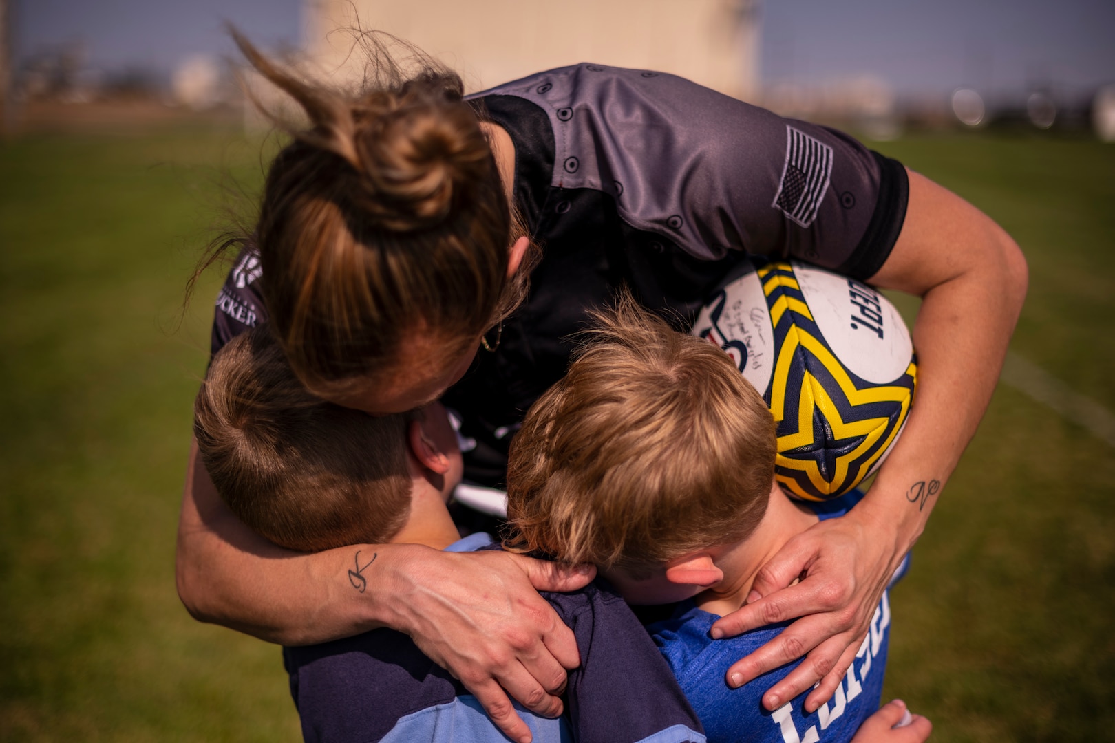 A standing woman holds her sons with her arms while a rugby ball is tucked away in her arms as well.