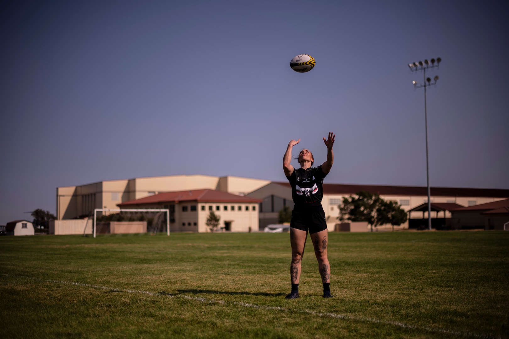 A woman stands on a soccer field and tosses a rugby ball in the air above her head.