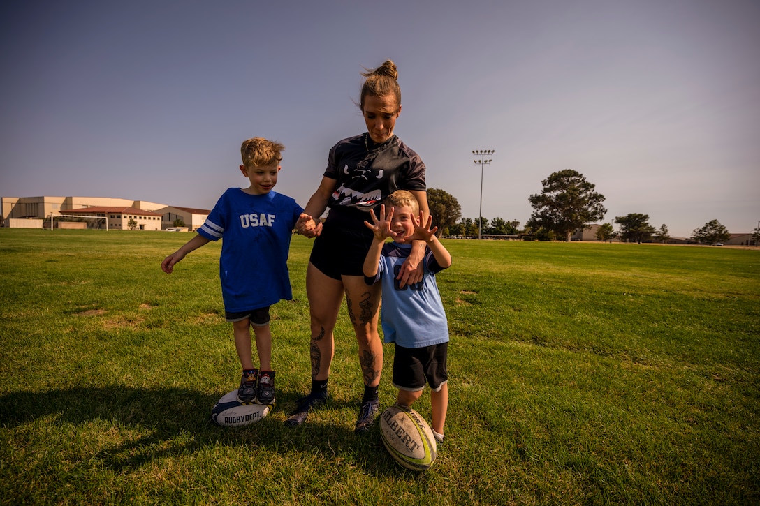 A woman stands with her two young boys on a soccer field while they hold rugby balls.