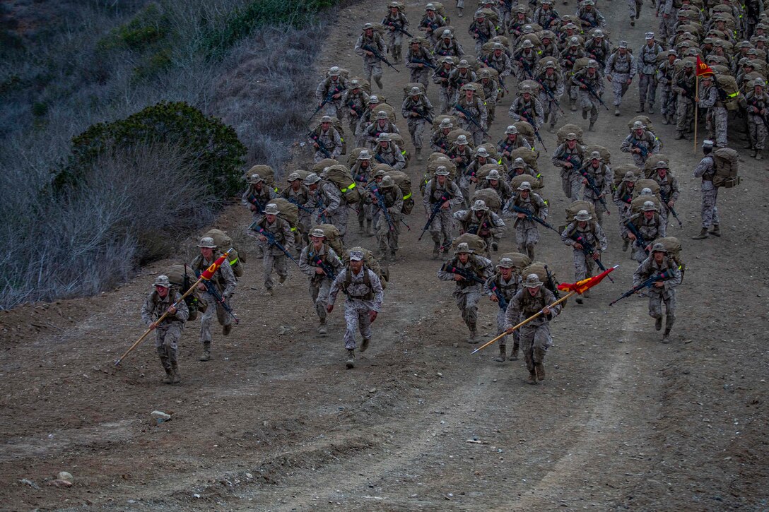 A large group of Marines carrying heavy packs run up a hill on a dirt road.