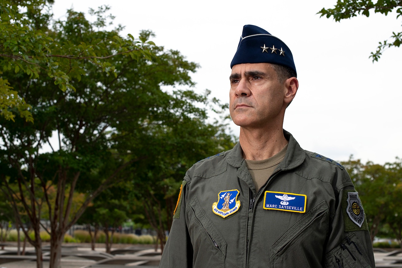 A man in a military uniform stands among trees and benches in a memorial area.