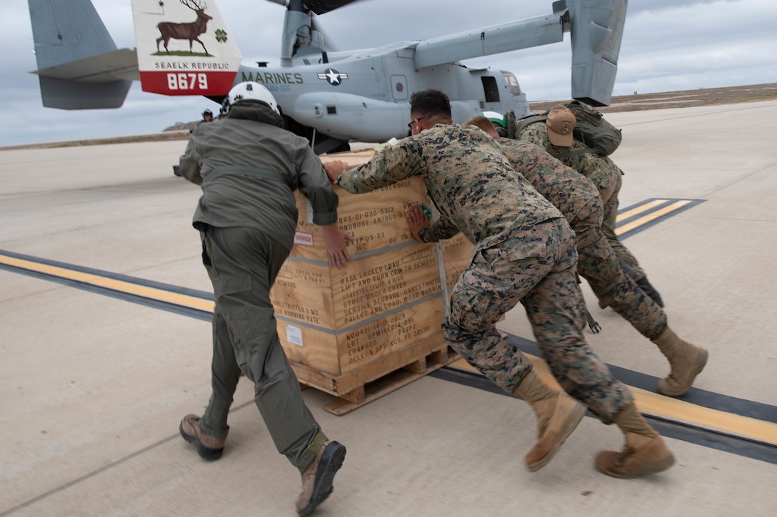 Sailors and Marines push a crate across the tarmac toward an aircraft.