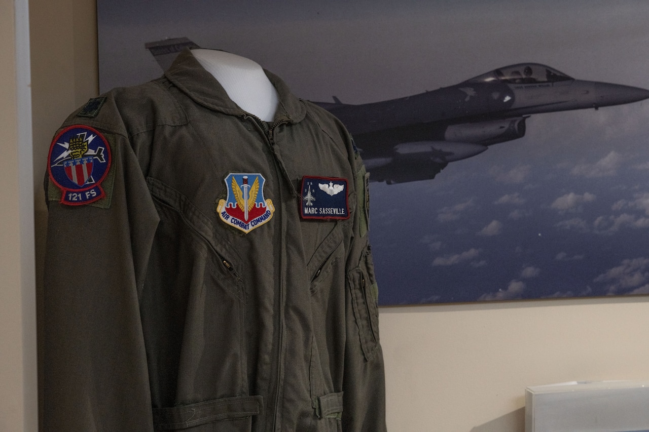 A military flight suit is displayed in front of a poster of an F-16.