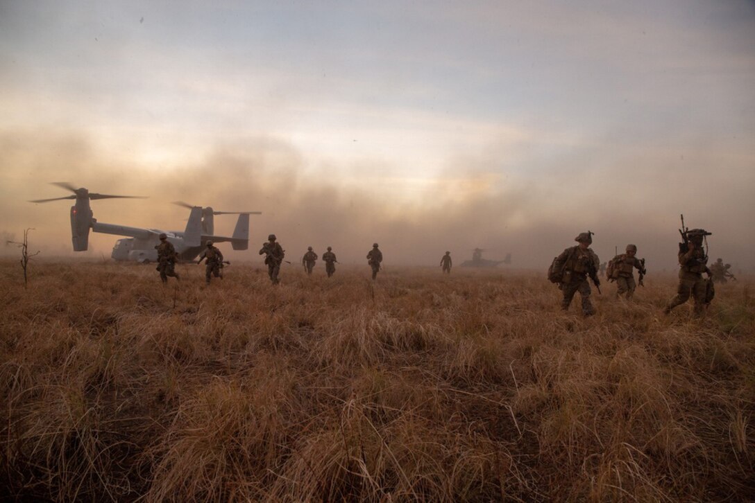 Service members walk through a field of long, brown grass with clouds of smoke behind them.