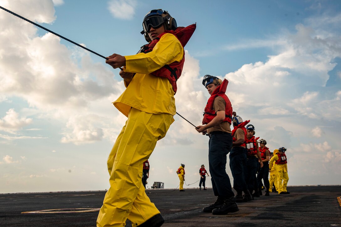 Sailors wearing protective gear pull a line while standing aboard a ship.