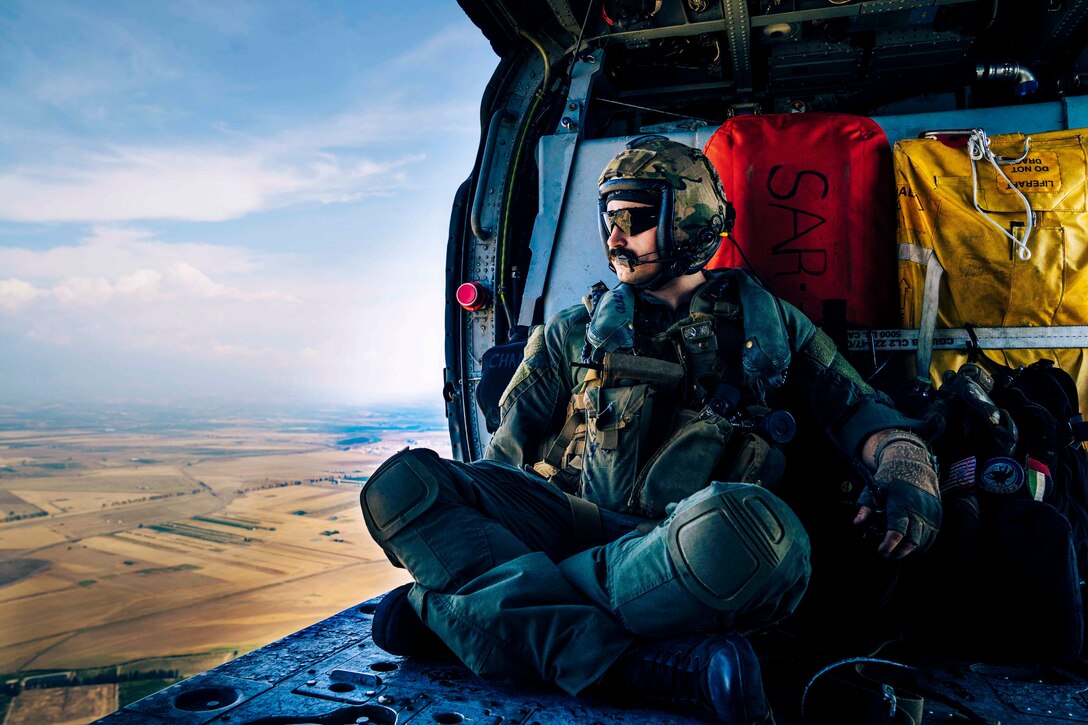 A sailor sits inside an airborne helicopter looking out through an open door.