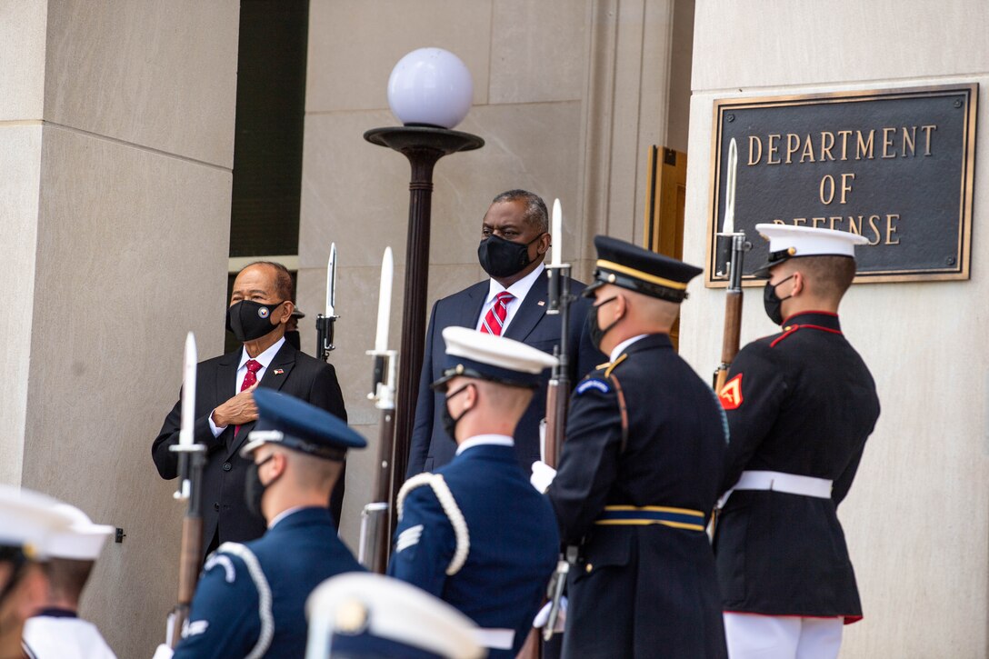Two men stand at attention. A military honor guard is in the foreground.