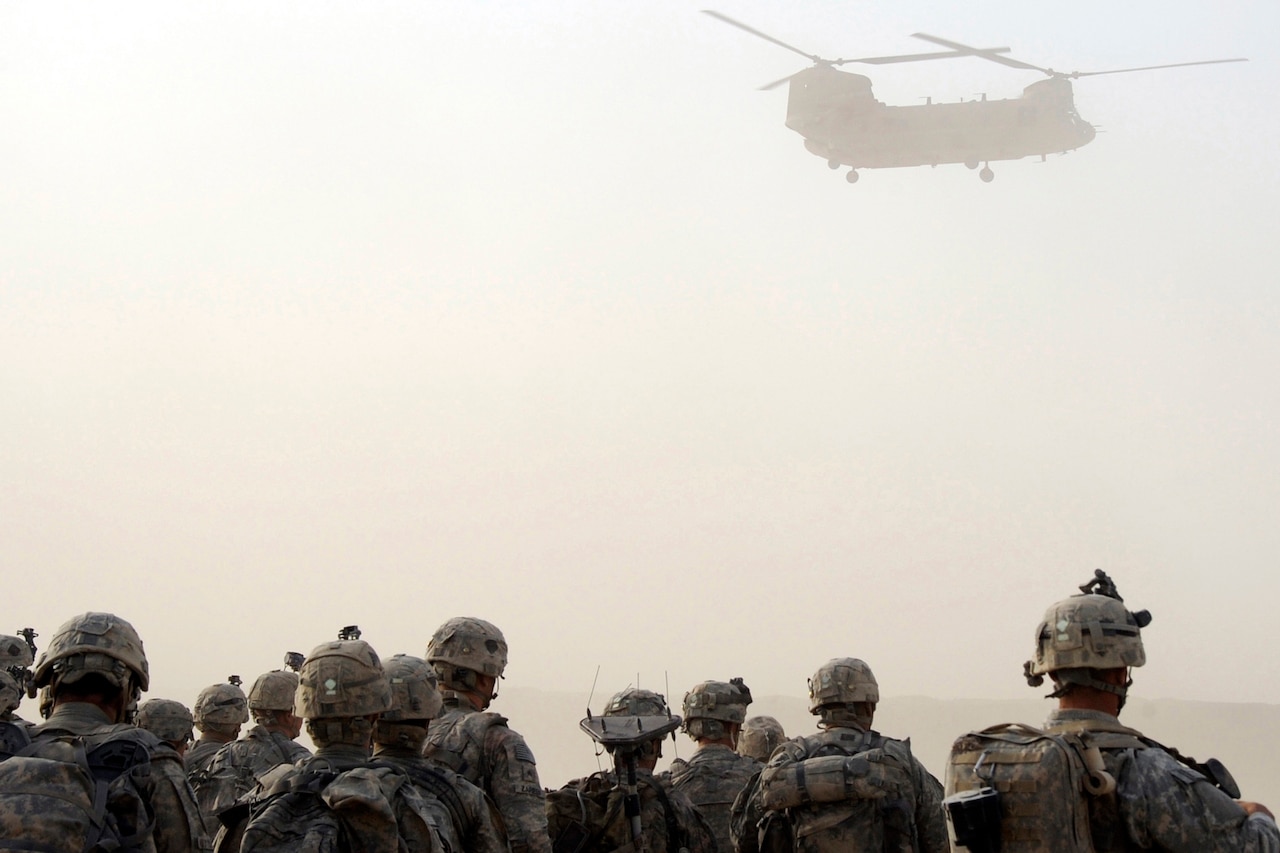 Soldiers stand on the ground beneath a hovering helicopter.