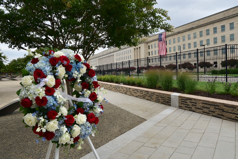 A wreath is placed on a stand. In the background, a flag is unfurled on a building.