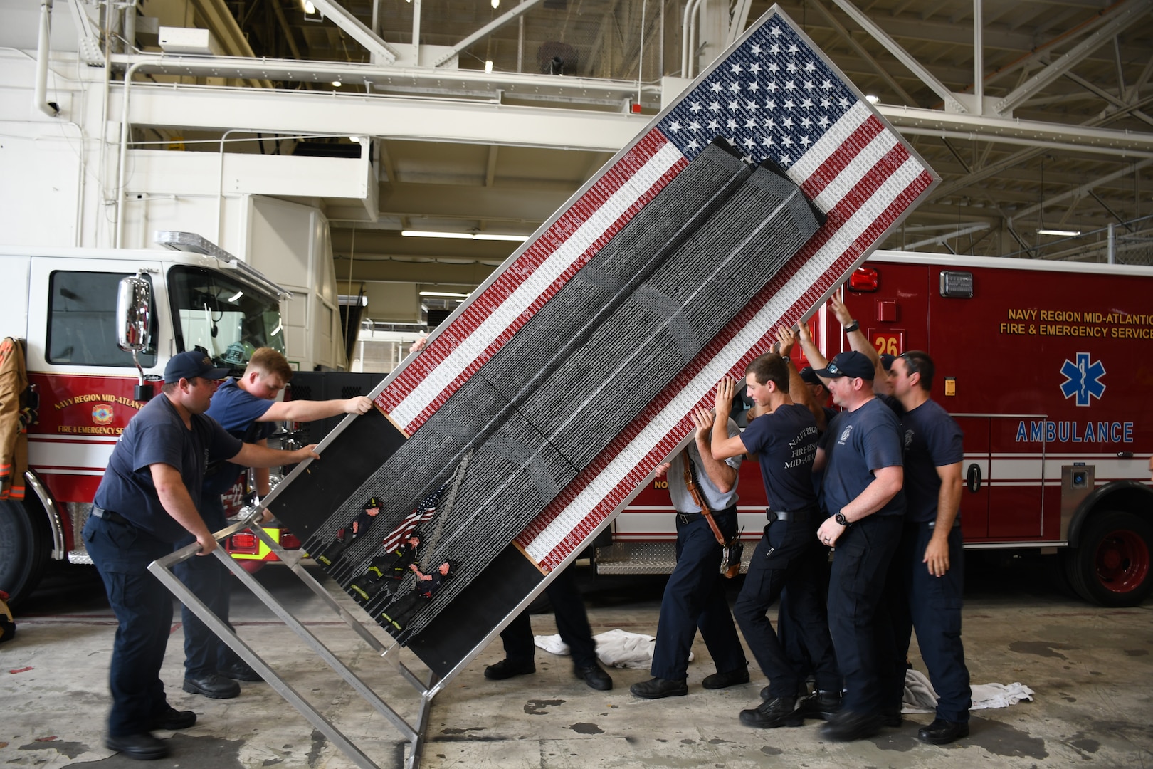 Members of the Navy Region Mid-Atlantic Fire and Emergency Services at Norfolk Naval Shipyard (NNSY) raise the 9/11 memorial artwork called “Divinity Among Heroes.” NNSY SurgeMain Sailor, Machinist Mate Chief Petty Officer (MMC)  Joseph Pisano, created the tribute piece during his off duty hours. The tribute piece is made up of 30,000+ drywall and trim screws, 2,978 one-inch wooden blocks, the base, a simulated fire truck, is constructed of authentic gauges, cranks, handles, hoses, chevrons, and plates from a decommissioned fire truck and it stands 12 foot tall by four foot wide.