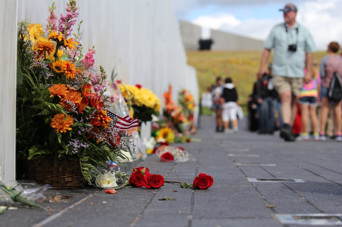 Visitors walk along a memorial wall adorned with flower bouquets.