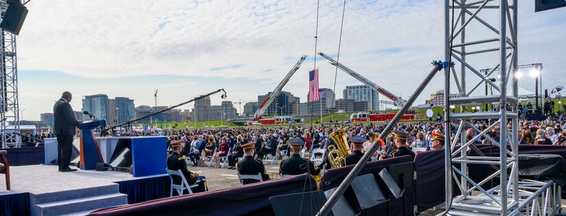 The secretary of defense stands at a lectern while speaking to a wide audience while fire trucks display the American flag in the backdrop.