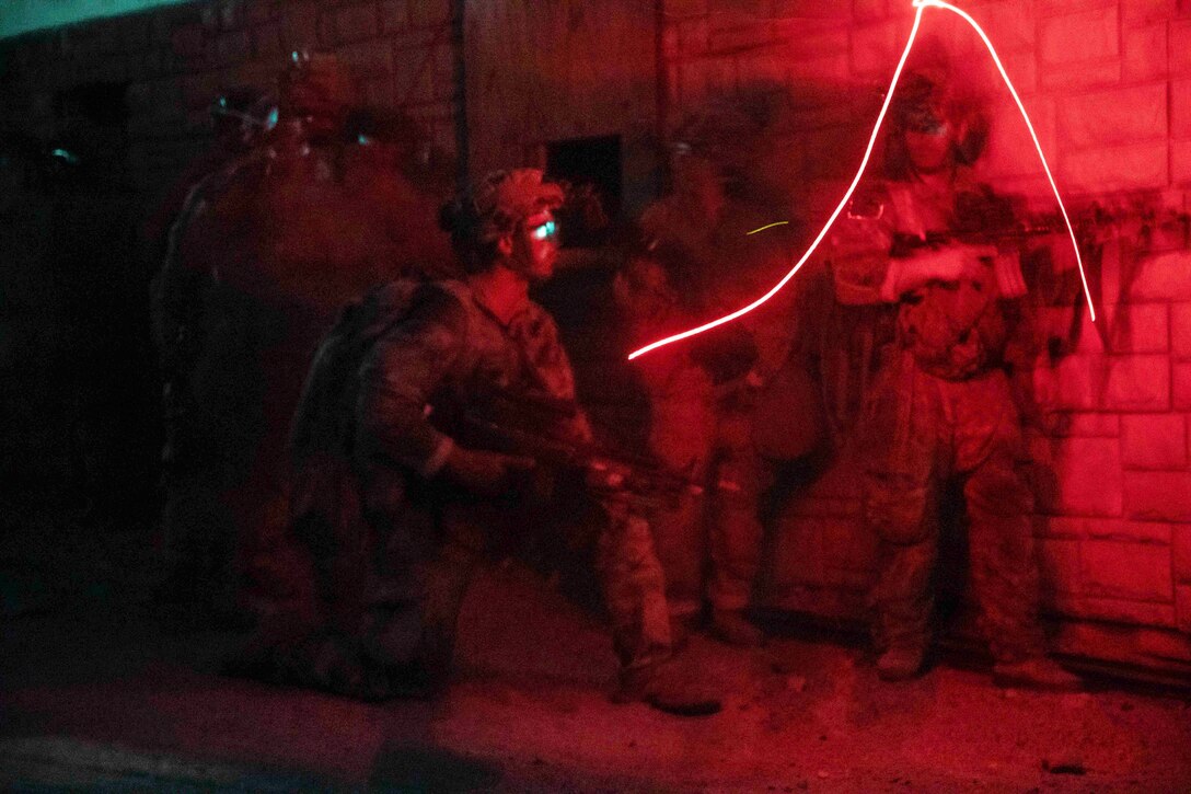 Soldiers hold weapons near a wall in the dark illuminated by red light.