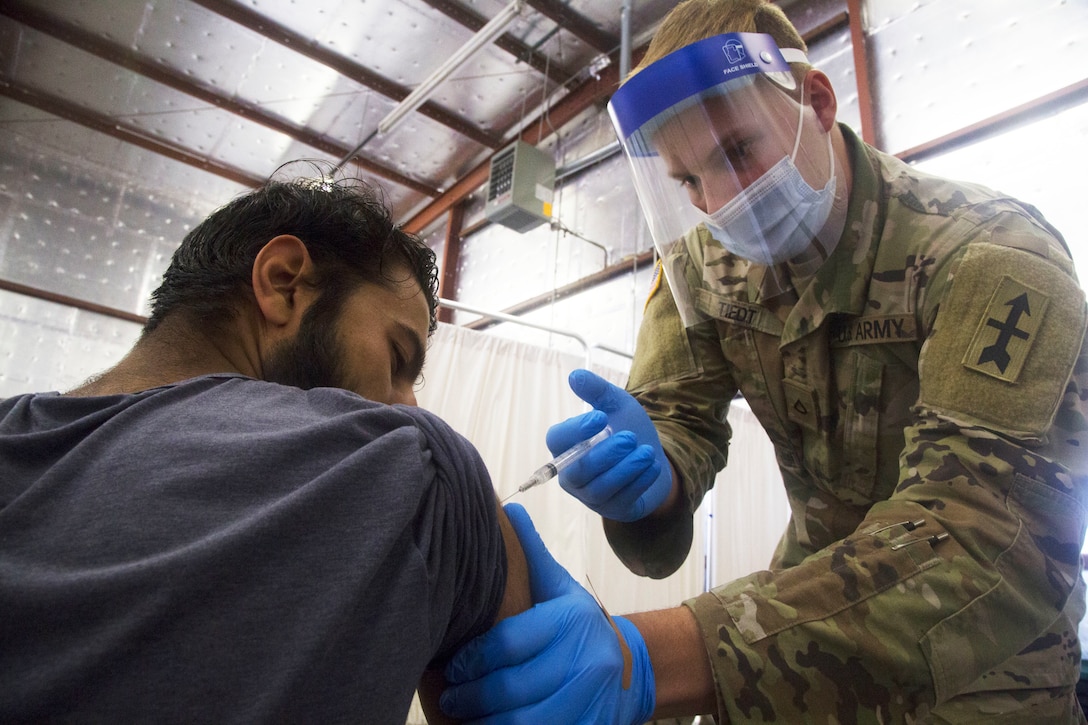 A soldier wearing a face mask and gloves bends over while holding a syringe and gives an Afghan evacuee sitting a COVID-19 vaccine.