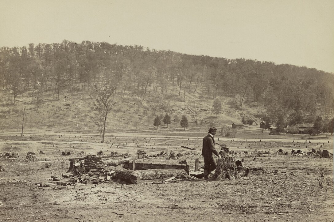 A man walks past debris in an open field. A wooded hill is in the background.