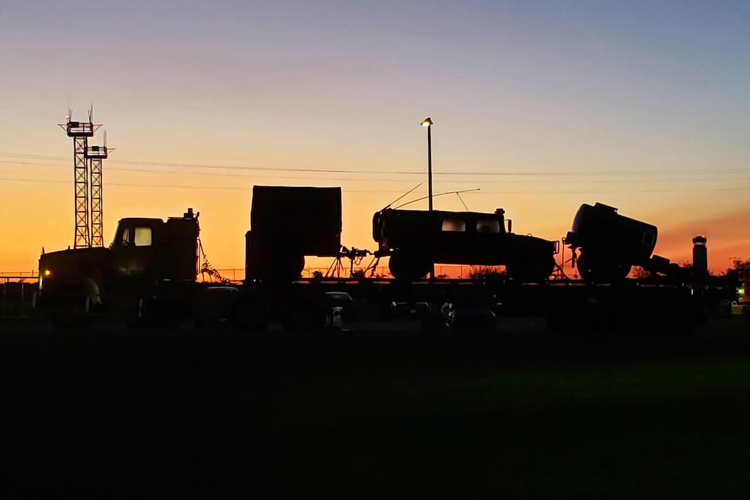 Vehicles and equipment sit on a tractor-trailer, shown in silhouette against a dawn sky.
