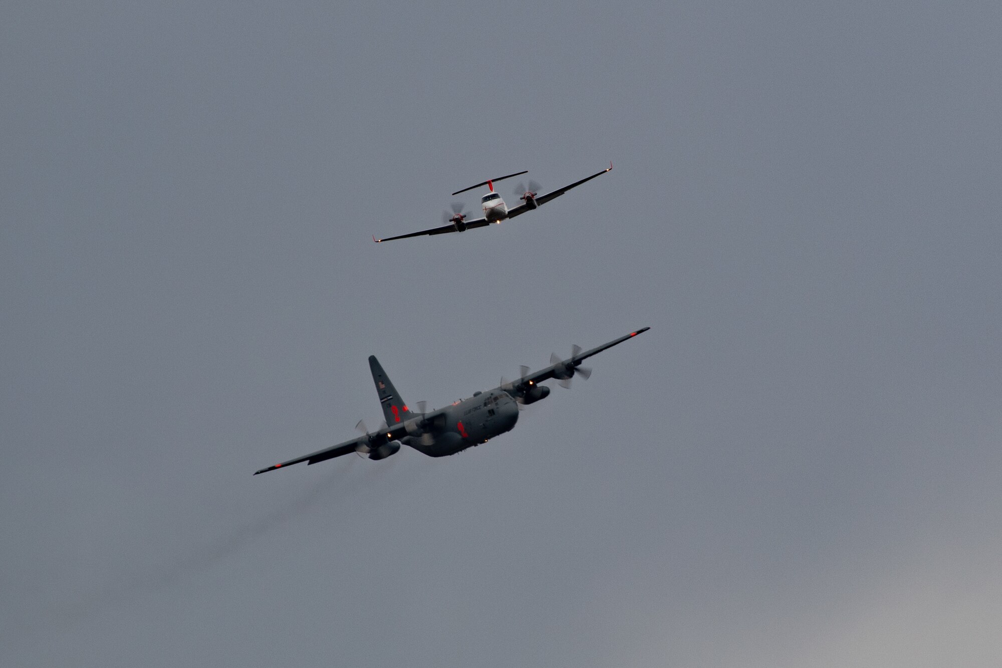 A C-130 marked with an orange 2 flies behind a smaller civilian plane.