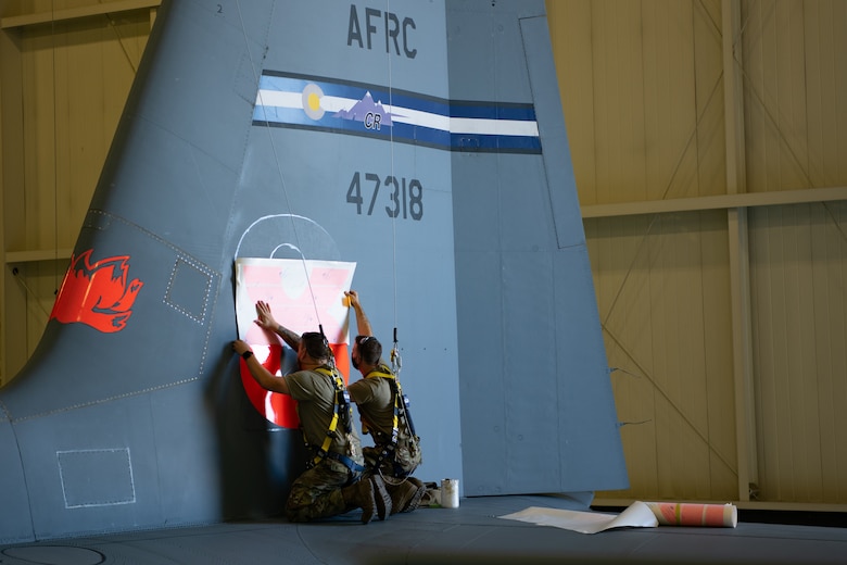 Two maintainers wearing fall restraint harnesses standing on the tail of a C-130 aircraft apply a large orange 8 to the aircraft.