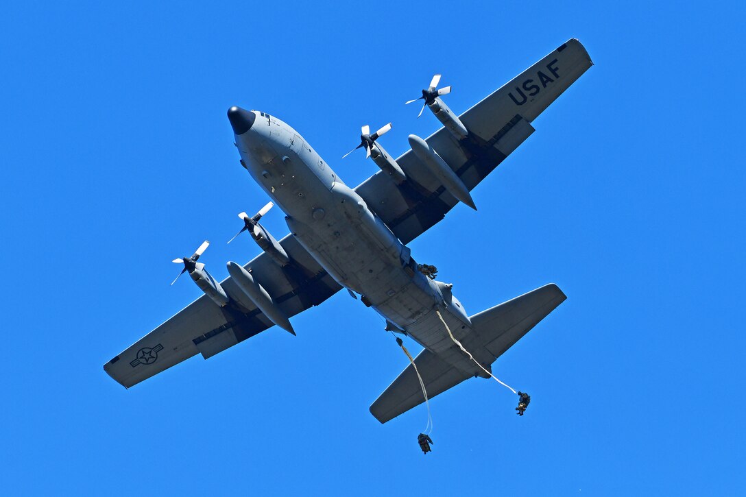 Several paratroopers hang from a large aircraft against a blue sky.