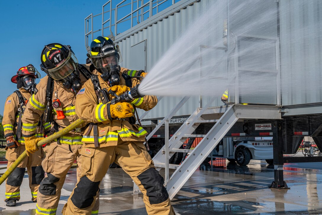 Three airmen in firefighting gear spray water from a hose.
