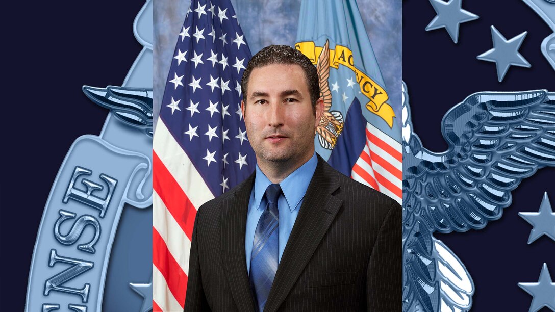 Official portrait of man sitting in front of American and DLA flags.