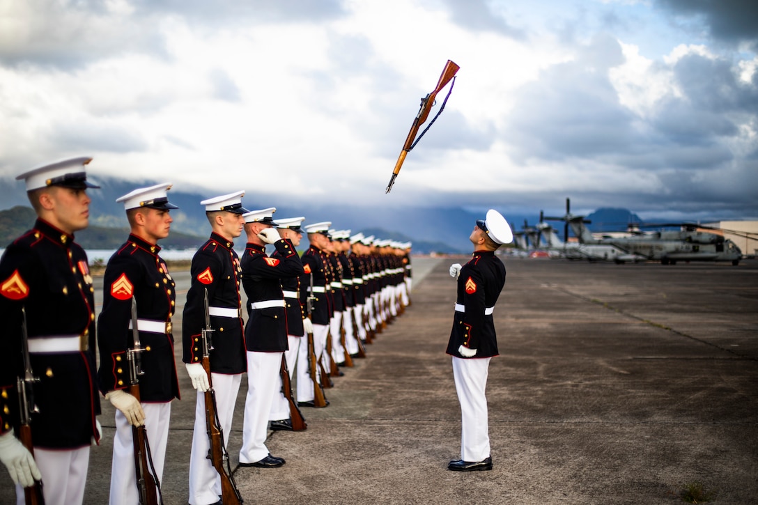 Marines in dress uniforms line up while a single Marine tosses his rifle in the air.
