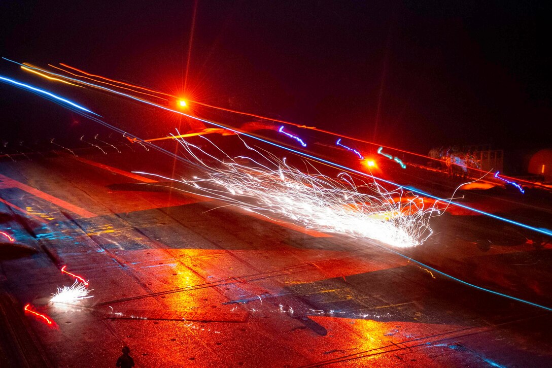 An aircraft lands on a ship at sea in the dark illuminated by colorful lights.