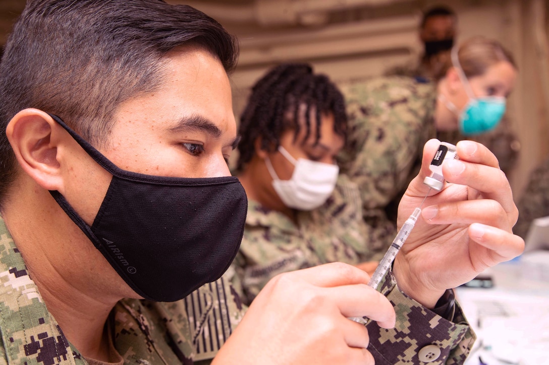 A sailor wearing a face mask holds a syringe while inserting the needle into a small bottle.