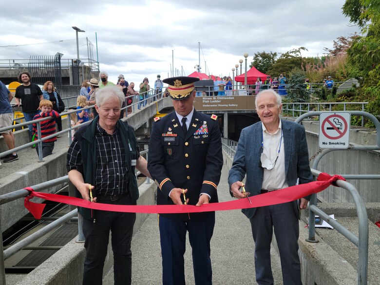 Photo of three men: one with grey hair is Jim Adams, the executive director of Discover Your Northwest, Col. Alexander 'Xander' Bullock, USACE Seattle district commander in the middle, and Rich Deline, director of The Corps Foundation on the right. cutting the ribbon to open the newly renovated fish ladder viewing room to the public, at Lake Washington Ship Canal and Hiram M. Chittenden Locks.