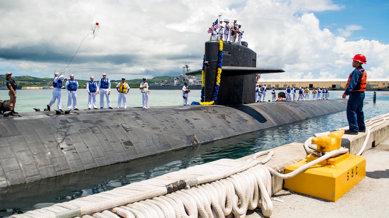Troops stand on submarine that is mooring pierside, where a sailor watches.