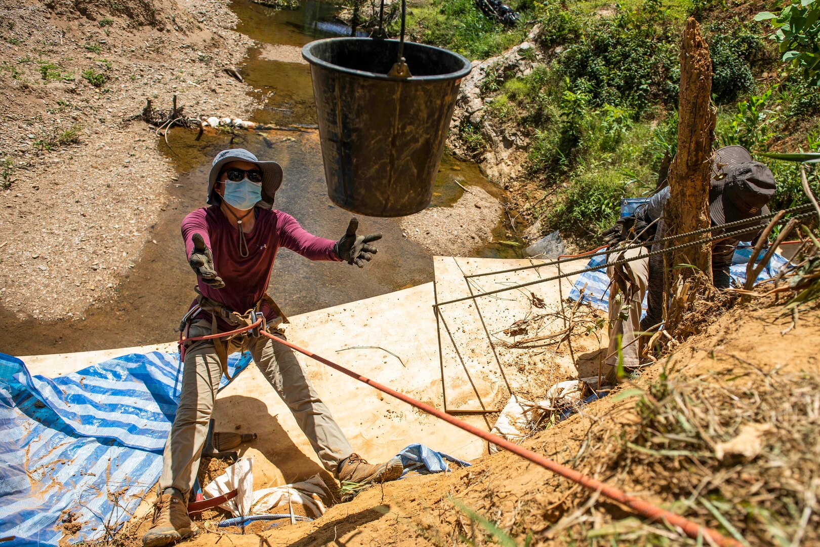 A bucket is lowered to a person suspended by a rope on the side of an embankment.