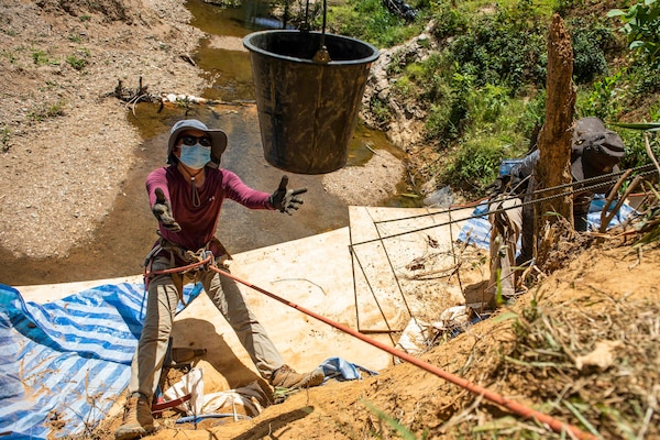 A bucket is lowered to a person suspended by a rope on the side of an embankment.