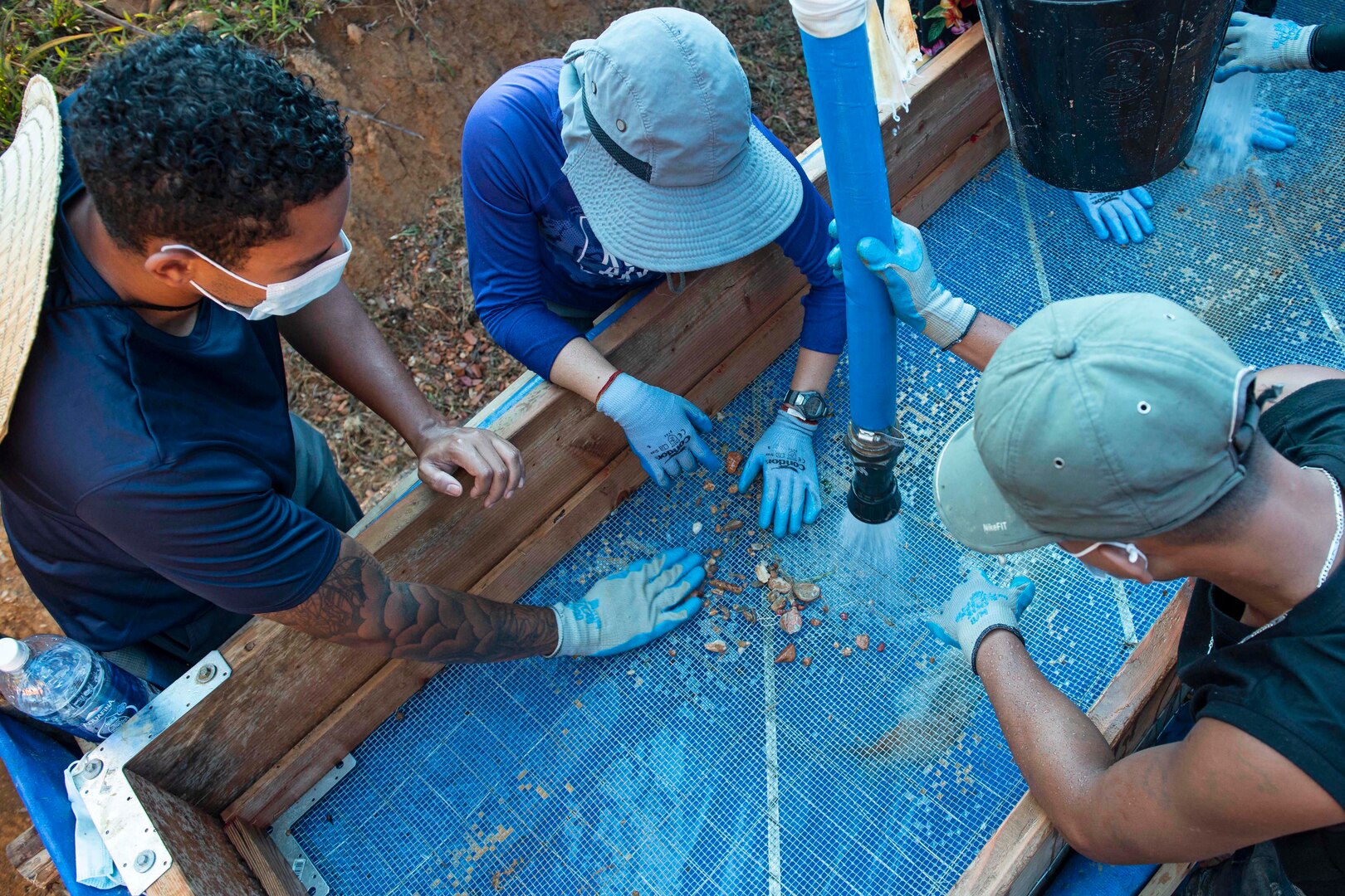 Three people look at debris in a large sieve as one person sprays it with a large hose.
