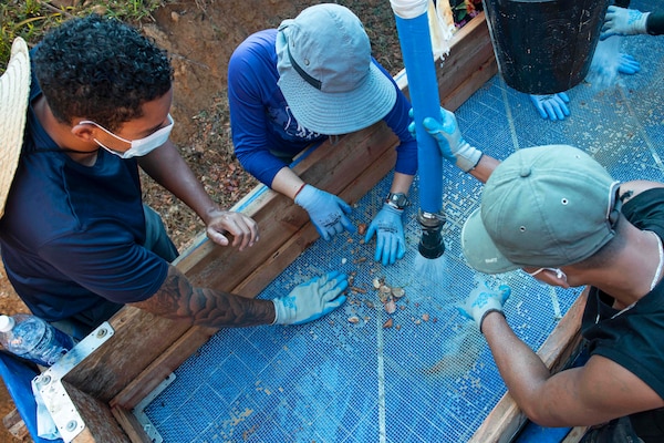 Three people look at debris in a large sieve as one person sprays it with a large hose.