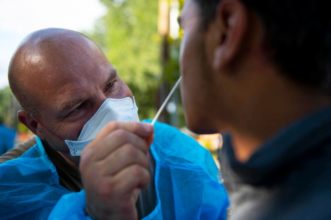 An airman wearing a face mask and medical gown holds a nasal swab up to a man's nose.