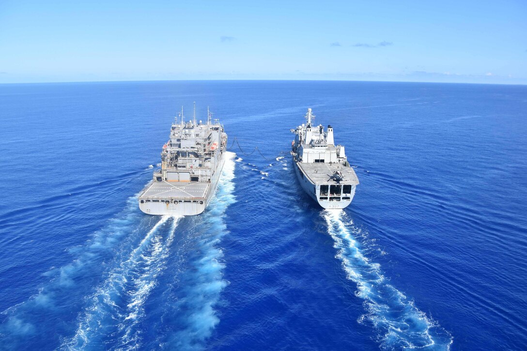 Two military ships sail next to each other with a hose connecting them.
