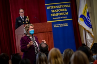 A woman wearing a blue face mask in a red blazer address a crowd in an auditorium, while another woman in uniform stands behind a podium. A banner in the background reads "International Seapower Symposium."