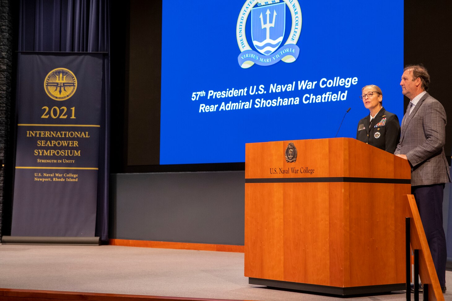 A woman in uniform speaks and stands next to a man in a suit behind a podium that reads "U.S. Naval War College."